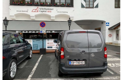 Behindertenparkplatz - Franz-Reisch-Straße 1 Eine graue Lieferwagen parkt vor einem Geschäft mit einem Schild „Schelhammers Capital 1832“. Im Hintergrund sind weitere Autos und ein Gebäude mit Fenstern zu sehen.
