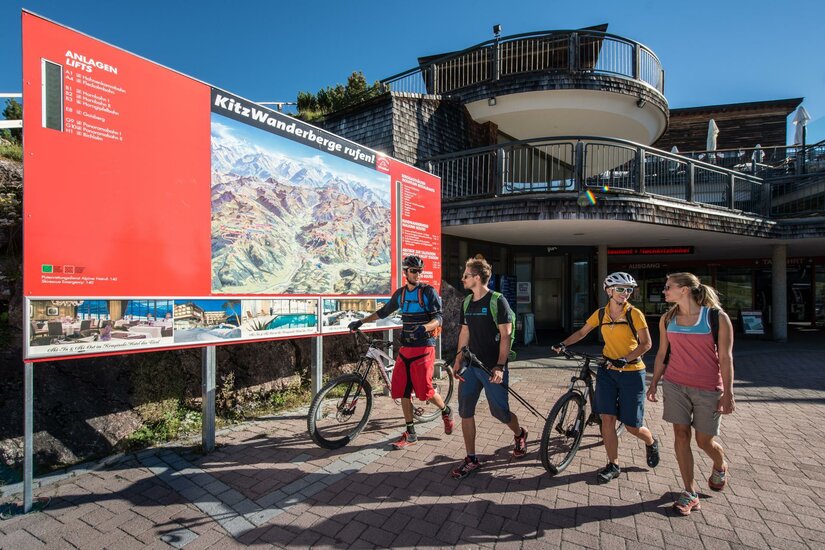 Bergbahn Kitzbühel © Kitzbühel Tourismus - Michael Eine Gruppe von Radfahrern steht vor einer Informationswand, die Wander- und Radwege zeigt. Im Hintergrund sind moderne Gebäude und strahlend blauer Himmel zu sehen.