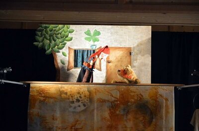 Eine Kasperl-Puppe und ein Stoffbär auf einer kleinen Theaterbühne mit gemaltem Hintergrund.