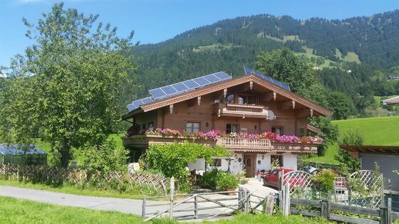 Haus Berger im Sommer Zweistöckiges Holzhaus mit Blumenschmuck am Balkon, Photovoltaikanlage auf dem Dach und Bergen im Hintergrund.