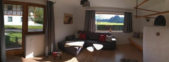 Ein gemütliches Wohnzimmer mit einem grauen Sofa und bunten Kissen. Durch das Fenster ist eine grüne Wiese und Berge im Hintergrund zu sehen.