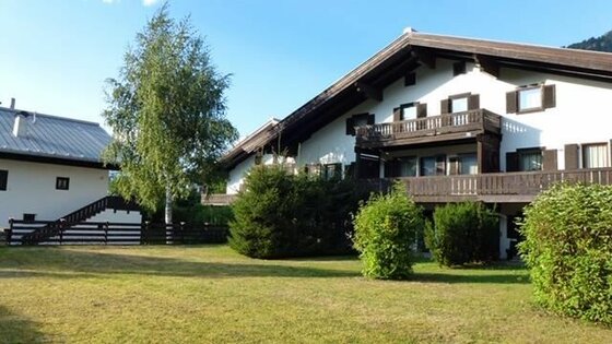Zweistöckiges Alpenhaus mit Holzbalkonen, weißer Fassade und angrenzender Rasenfläche bei Tageslicht.