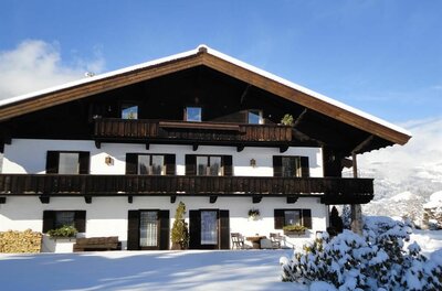 Landhaus Feller im Winter Zweistöckiges Landhaus mit Holzbalkonen und weißer Fassade in verschneiter Landschaft unter blauem Himmel.