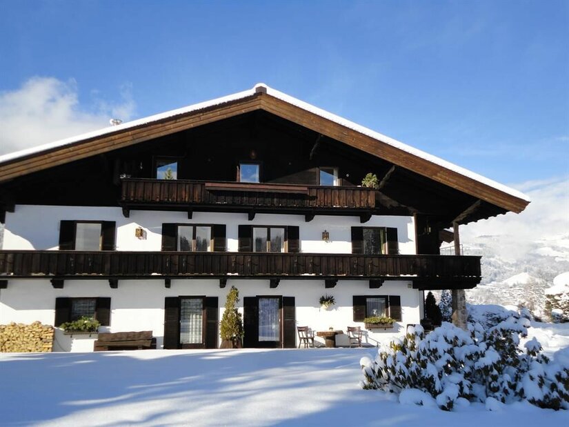 Zweistöckiges Landhaus mit Holzbalkonen und weißer Fassade in verschneiter Landschaft unter blauem Himmel.