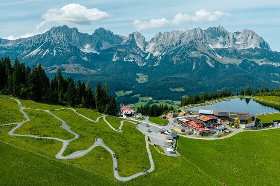 Going_PonyAlm_Panorama_Wilder Kaiser Eine malerische Landschaft mit grünen Wiesen und majestätischen Bergen im Hintergrund. Im Vordergrund liegt eine kleine Siedlung mit einem Teich und gewundenen Wegen.