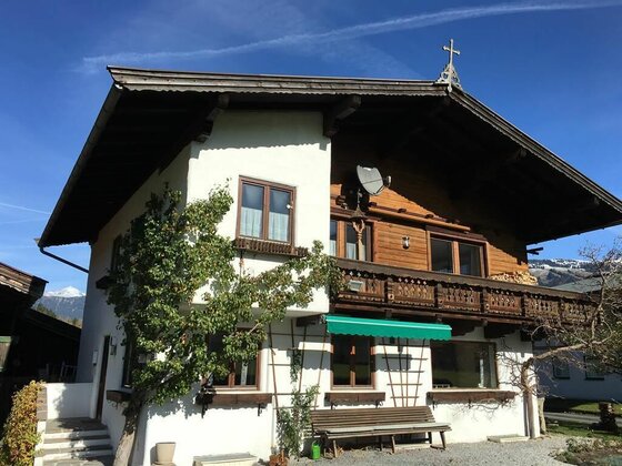 Zweistöckiges Haus mit weißer Fassade, Holzbalkon und grünem Sonnenschutz unter blauem Himmel.