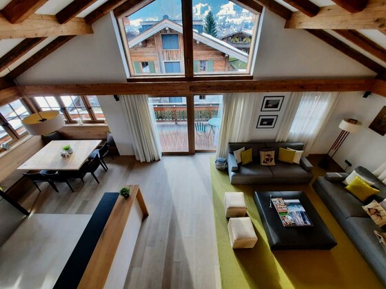 Moderner Wohnbereich mit Holzbalken, Sofas, Esstisch und großem Giebelfenster mit Blick auf Chalets.