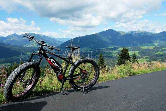 E-Fatbike am Kitzbüheler Horn Ein schwarzes E-Fatbike steht auf einer asphaltierten Straße vor einem Bergpanorama.