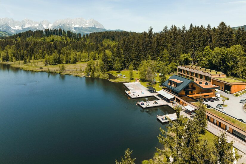 Luftaufnahme eines Holzgebäudes mit Badestegen an einem See vor Wald und Gebirgskette.