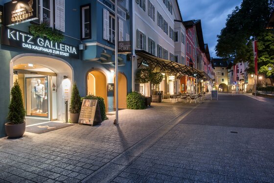 Geschäfte Innenstadt Sommer Gepflasterte Fußgängerzone in Kitzbühel mit beleuchteten Schaufenstern und historischen Fassaden in der Dämmerung.