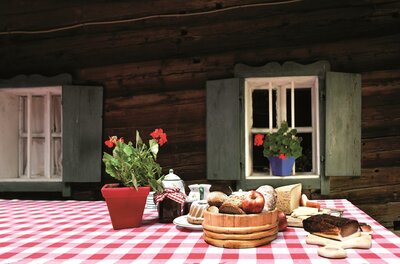 Achrainhof - Symbolfoto <p>Ein Tisch mit rot-weißen Tischdecken, auf dem verschiedene Lebensmittel und eine Blume stehen. Im Hintergrund sieht man Holzwände und Fenster mit grünen Fensterläden.</p>