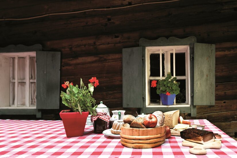 Achrainhof - Symbolfoto <p>Ein Tisch mit rot-weißen Tischdecken, auf dem verschiedene Lebensmittel und eine Blume stehen. Im Hintergrund sieht man Holzwände und Fenster mit grünen Fensterläden.</p>