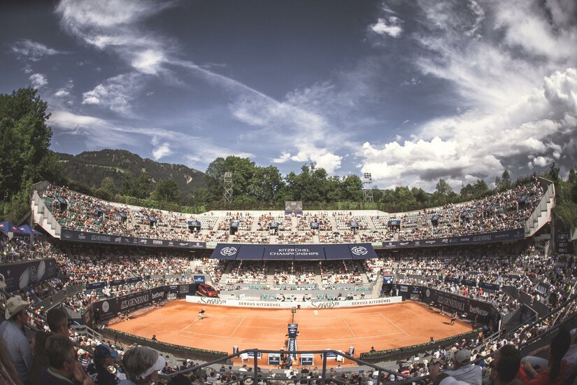 Tennisstadion Ein Tennisplatz mit roten Sand und vielen Zuschauern auf den Tribünen. Der Himmel ist klar und es sind einige Wolken zu sehen.