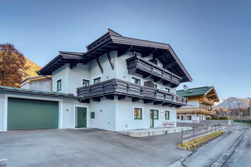 Außenansicht NA-Home Kitzbühel Modernes alpines Wohnhaus mit weißer Fassade, dunklen Holzbalkonen und einer Garage an einer asphaltierten Straße.