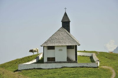 Eine kleine Kirche mit einem spitzen Dach steht auf einem Hügel. Im Hintergrund grasen Kühe auf der grünen Wiese.