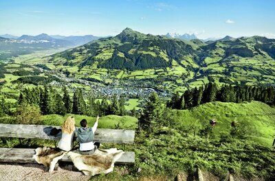 Aussichtspunkt am Hahnenkamm Zwei Personen sitzen auf einer Holzbank und blicken auf ein Tal mit Bergen im Hintergrund.