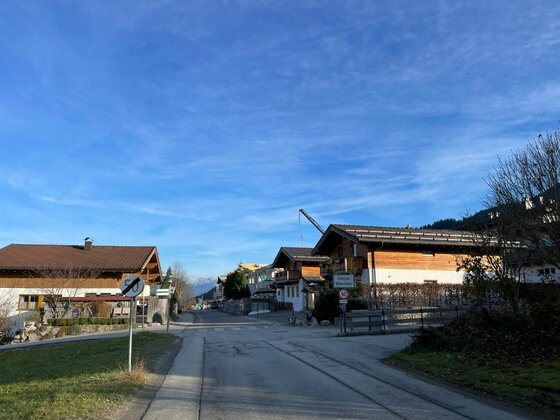 Radweg Wilder Kaiser Asphaltierte Straße führt durch eine Siedlung mit alpinen Holzhäusern unter blauem Himmel.