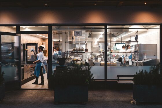 Einblick in die Wirtshausküche Blick durch eine große Glasfront in eine hell erleuchtete, moderne Restaurantküche mit arbeitenden Köchen.