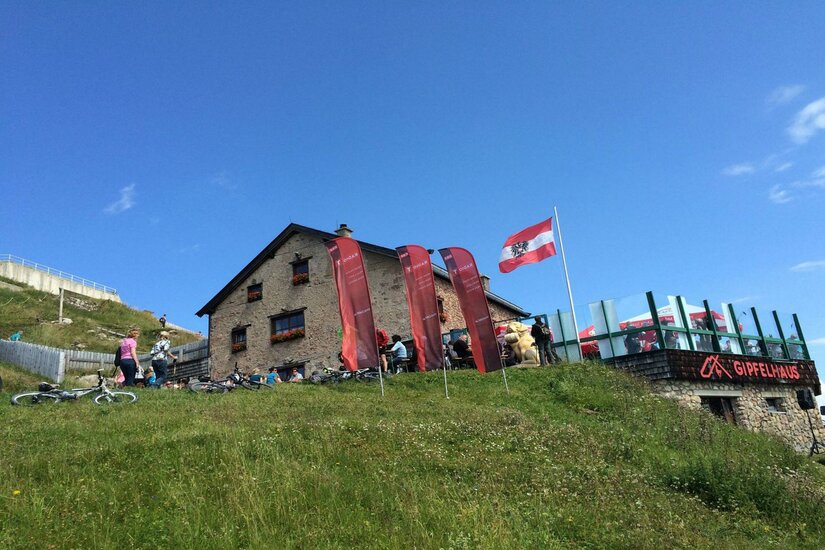 Ein gemütliches Berghaus mit einer Terrasse, umgeben von Wiese und grünen Hügeln. Einige Personen sind unterwegs, und eine Fahne weht im Wind.