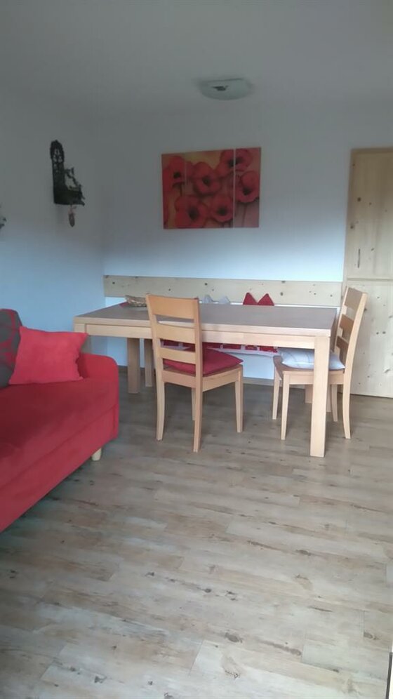 Wohnzimmer mit hellem Holztisch, zwei Stühlen, einer Eckbank und einem roten Sofa.