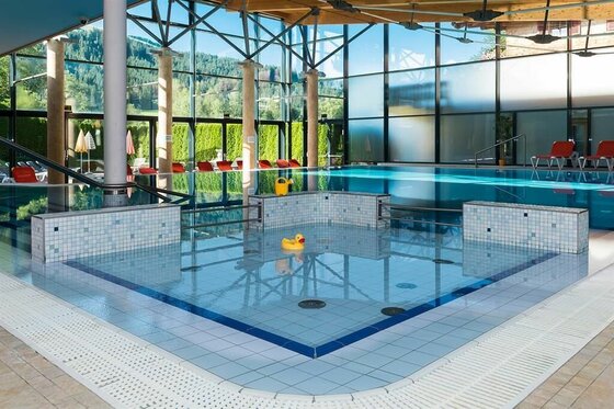 Innenansicht eines Hallenbads mit Kinderbecken, großem Schwimmbecken und Glasfront mit Blick auf bewaldete Berge.