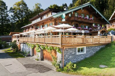 Mehrstöckiger Gasthof aus dunklem Holz mit großer Sonnenterrasse und weißen Schirmen.
