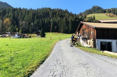 Ein Mountainbiker auf einer asphaltierten Straße neben einem Holzstadel vor bewaldeten Bergen.