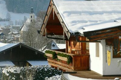 Aussicht vom Apartment Alpenland Verschneites Holzhaus mit Balkon vor einer Dorfkirche im Winter.