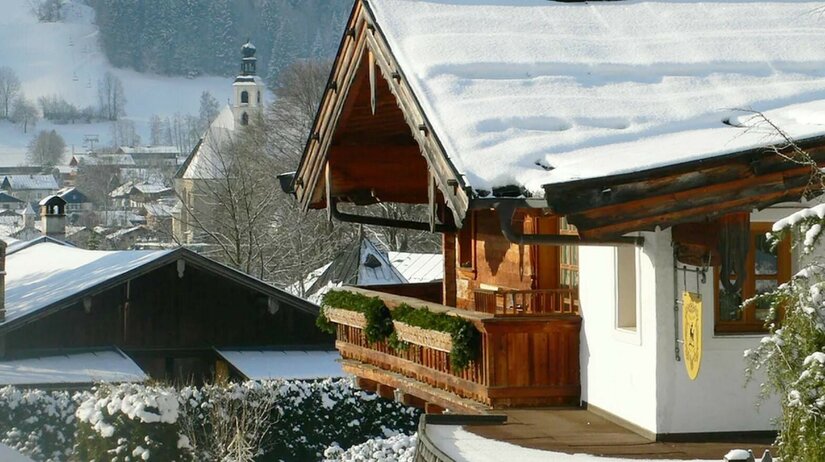 Aussicht vom Apartment Alpenland Verschneites Holzhaus mit Balkon vor einer Dorfkirche im Winter.