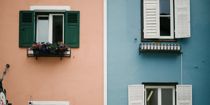 Zwei benachbarte Fensterfronten: eine in sanftem Rosa mit grünen Fensterläden und eine in beruhigendem Blau mit weißen Läden.