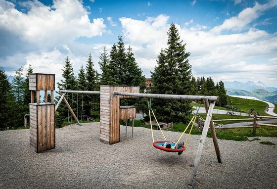 Ein Kind liegt in einer Nestschaukel auf einem Holzspielplatz vor Nadelbäumen und Bergen.