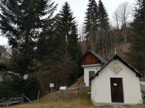 Kapelle am Kelchalmgraben Kleine weiße Kapelle mit Holztür und Wandgemälde vor Nadelbäumen am Kelchalmgraben.