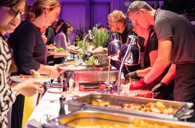 Ein Buffet mit verschiedenen Gerichten wird von mehreren Personen präsentiert. Gäste wählen aus einer Vielzahl von Speisen und genießen das Essen in einer festlichen Atmosphäre.