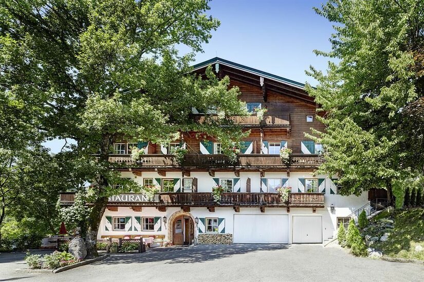 Landsitz Römerhof Außenansicht Mehrstöckiges alpines Gebäude mit Holzfassade, Balkonen und grün-weißen Fensterläden zwischen Bäumen.