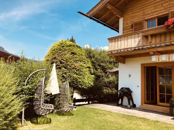 Alpines Holzhaus mit Garten, zwei Hängesesseln auf einer Wiese und einem Grill auf der Terrasse.