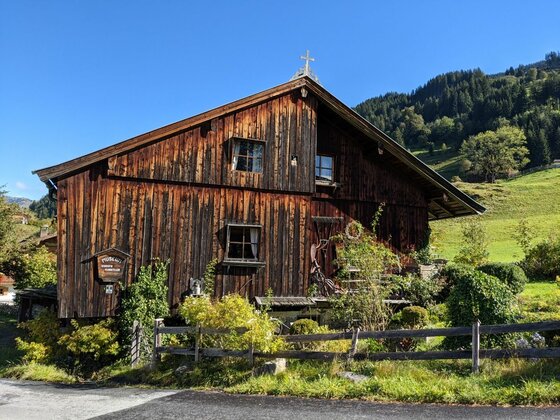 Holzhaus am Jochberger Wasserweg Rustikales Holzhaus mit dunkler Fassade vor bewaldetem Berghang unter blauem Himmel.