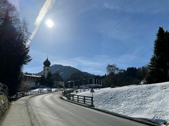 Asphaltierte Straße führt an einer Kirche mit Zwiebelturm und schneebedeckten Hängen vorbei.