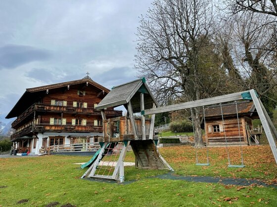 Holzspielplatz mit Kletterturm und Schaukel vor einem traditionellen Alpenhotel aus dunklem Holz.