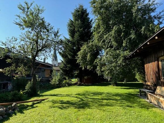 Ein ruhiger Garten mit grünem Rasen und hohen Bäumen. Im Hintergrund ist ein Kirchturm sichtbar.