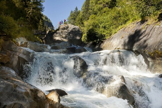 Das Bild zeigt die Umbalfälle mit mehreren Wasserstufen, über die das Wasser zwischen großen Felsen talwärts fließt. Links oberhalb des Wasserfalls ist eine Aussichtsplattform zu sehen, auf der mehrere Personen stehen. Die Felswände, das fließende Wasser und die dichte Vegetation prägen die Landschaft rund um den Wasserfall.