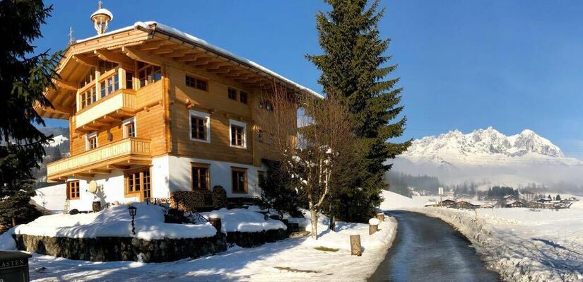 Haus Lazar im Winter Mehrstöckiges Holzhaus in verschneiter Landschaft mit Blick auf ein Gebirgsmassiv unter blauem Himmel.