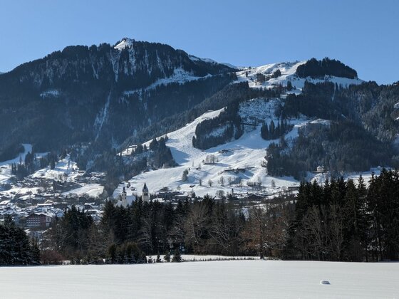 Winteransicht Kitzbüheler Alpen Verschneite Berglandschaft mit bewaldeten Hängen und einer kleinen Ortschaft im Tal unter blauem Himmel.