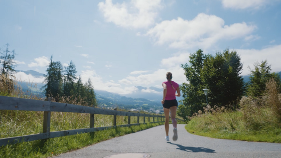 Laufen mit Lisa Hauser Eine Frau in Sportkleidung joggt auf einem asphaltierten Weg entlang eines Holzzauns in den Bergen.