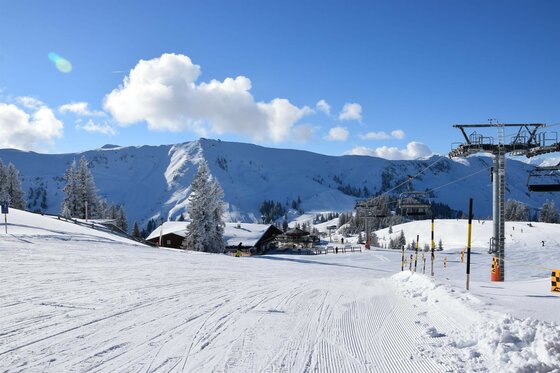 Sonnenrast im Winter Präparierte Skipiste mit Sessellift, Berghütte und verschneiten Bergen unter blauem Himmel.