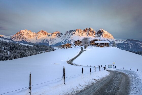 Bio Yoga Hof Kitzbühel Verschneite Berglandschaft mit einem Bauernhof und einer kurvigen Straße vor einem Gebirgsmassiv.