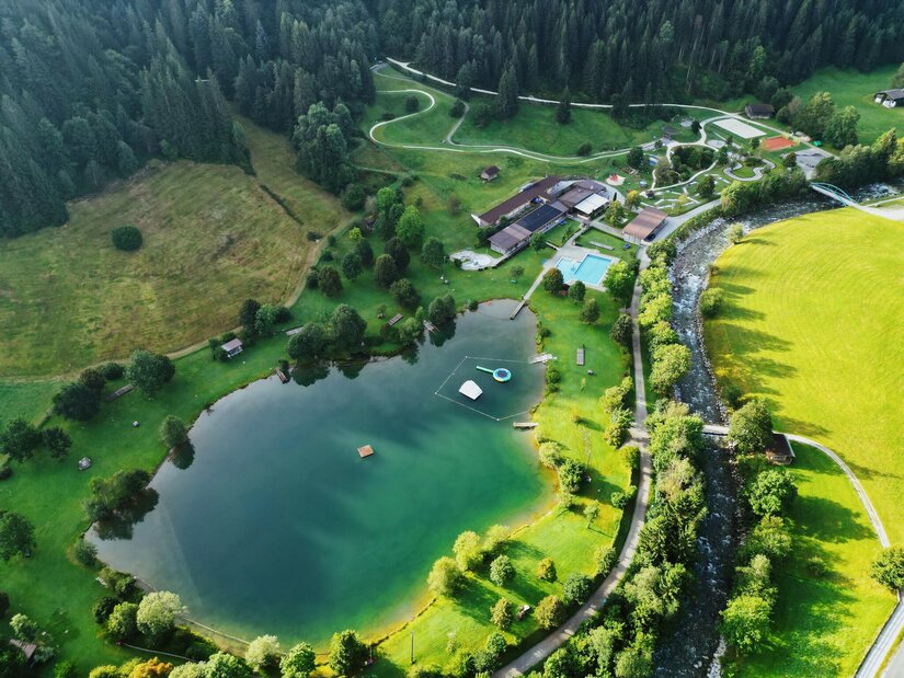 Badesee @ Region Hohe Salve-Magdalena Lainminger Eine malerische Landschaft mit einem See, umgeben von grünen Wiesen und Bäumen. Im Hintergrund sind Gebäude und ein Schwimmbad sichtbar.