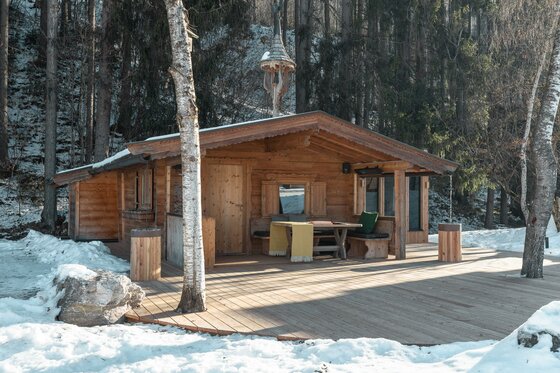 WEB_HansFrauenschuh_-64 Eine gemütliche Holzhütte inmitten einer verschneiten Waldlandschaft. Auf der Terrasse stehen Tische und Stühle, um die Aussicht zu genießen.