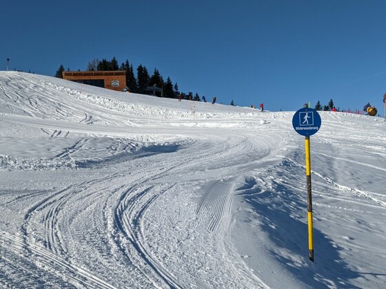 Winterwanderweg bei St. Bernhard Präparierter Winterwanderweg im Schnee mit blauem Hinweisschild vor blauem Himmel.