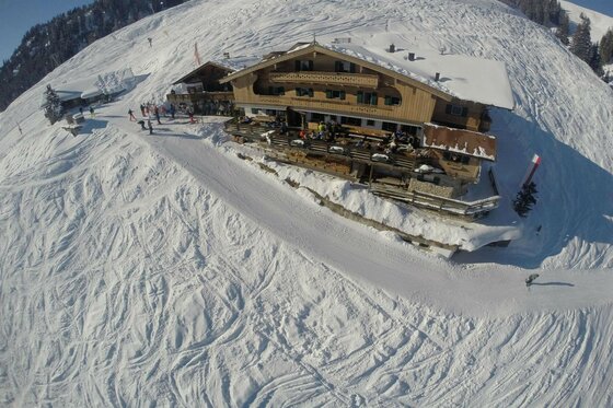 Melkalm im Winter Luftaufnahme einer schneebedeckten Almhütte aus Holz an einem Skihang mit Skifahrern.