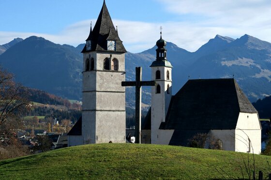 Kirche am Lebenberg Weiße Kirche mit zwei Türmen und großem Holzkreuz vor einer Gebirgskette.